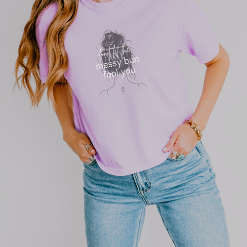 A woman wearing a light purple graphic t-shirt with a black line art illustration of a messy bun and the text 'Don't let  the messy bun fool you'. She is also wearing light wash blue jeans against a solid light gray background.