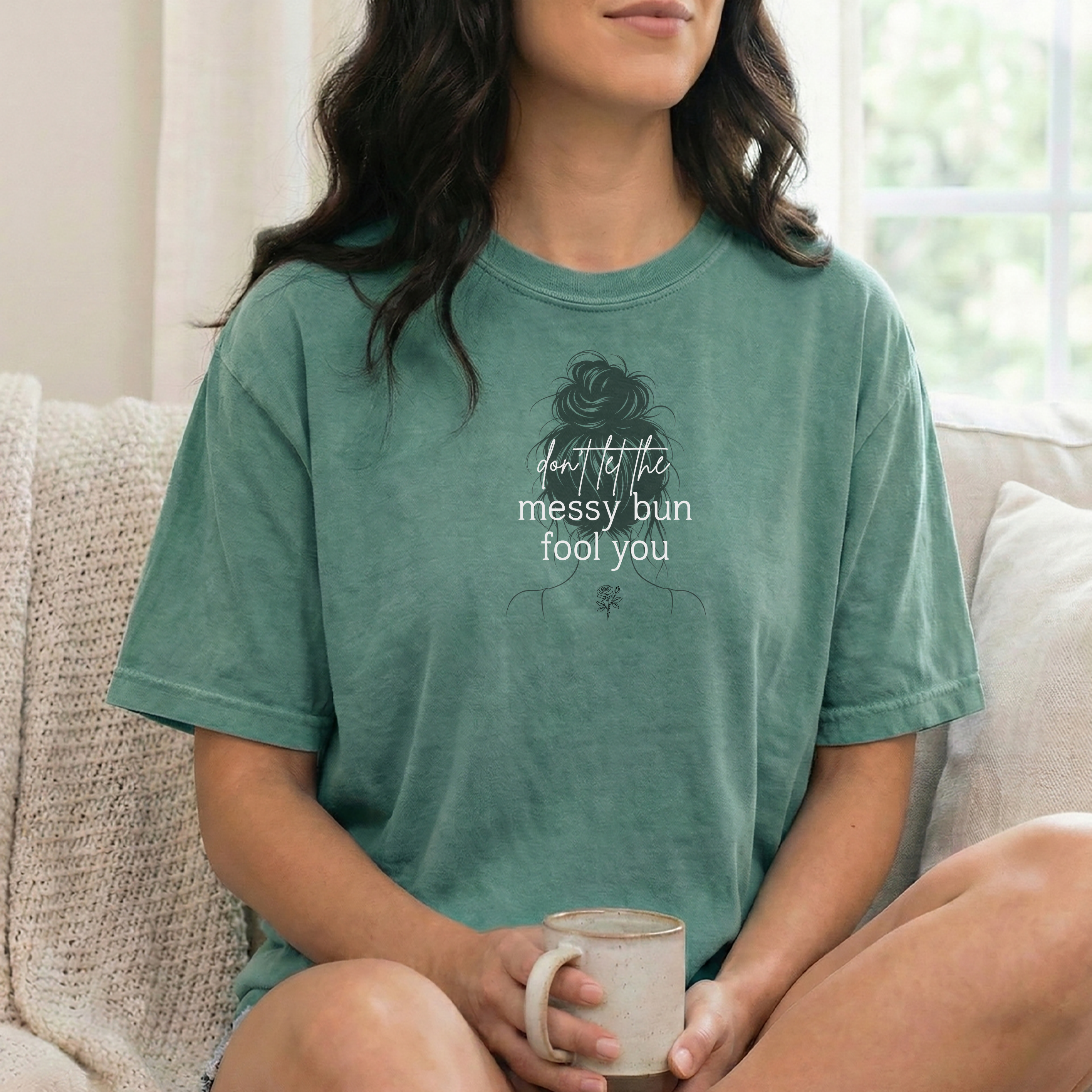 A woman wearing a light green short-sleeve graphic t-shirt with a silouette of a woman with a messy bun illustration and the text 'don't let the messy bun fool you' in white script sitting on a couch holding a coffee mug. 