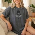 A woman wearing a dark grey short-sleeve graphic t-shirt with a silouette of a woman with a messy bun illustration and the text 'don't let the messy bun fool you' in white script sitting on a couch. 